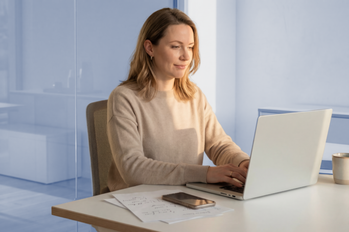 woman working at desk with her laptop and cellphone beside her. She looks happy and there is a neutral blue background.